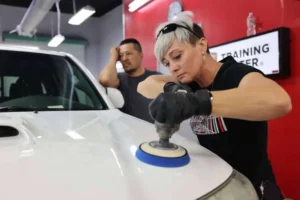 Woman using a buffer to polish a car hood while a man observes.