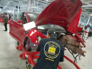 Auto technician working on the body of a red car with the hood open in an industrial setting.